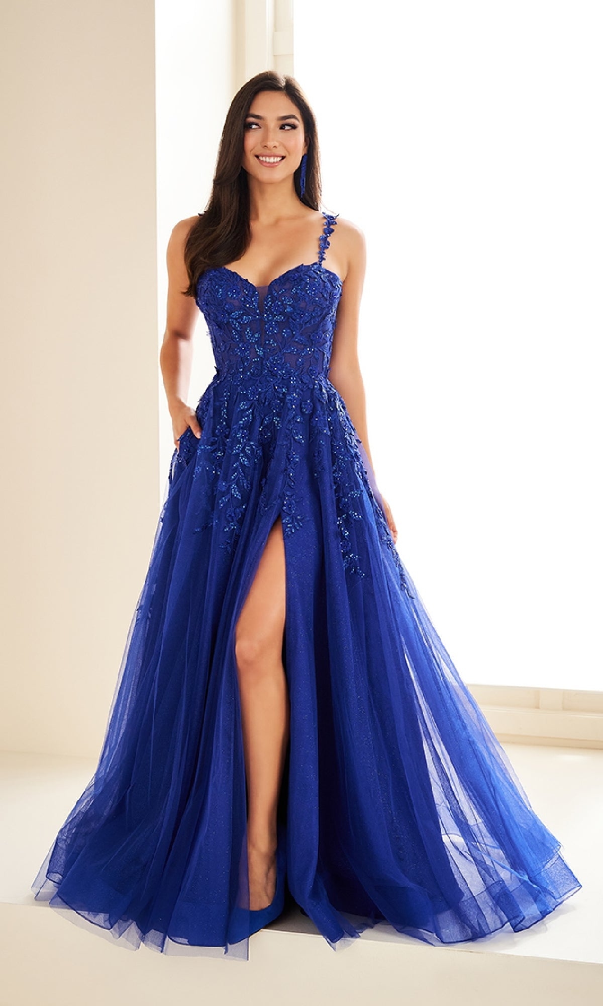 A woman smiles indoors wearing the Ellie Wilde EW36046, a sleeveless, floor-length royal blue prom dress featuring floral embellishments and a thigh-high slit.